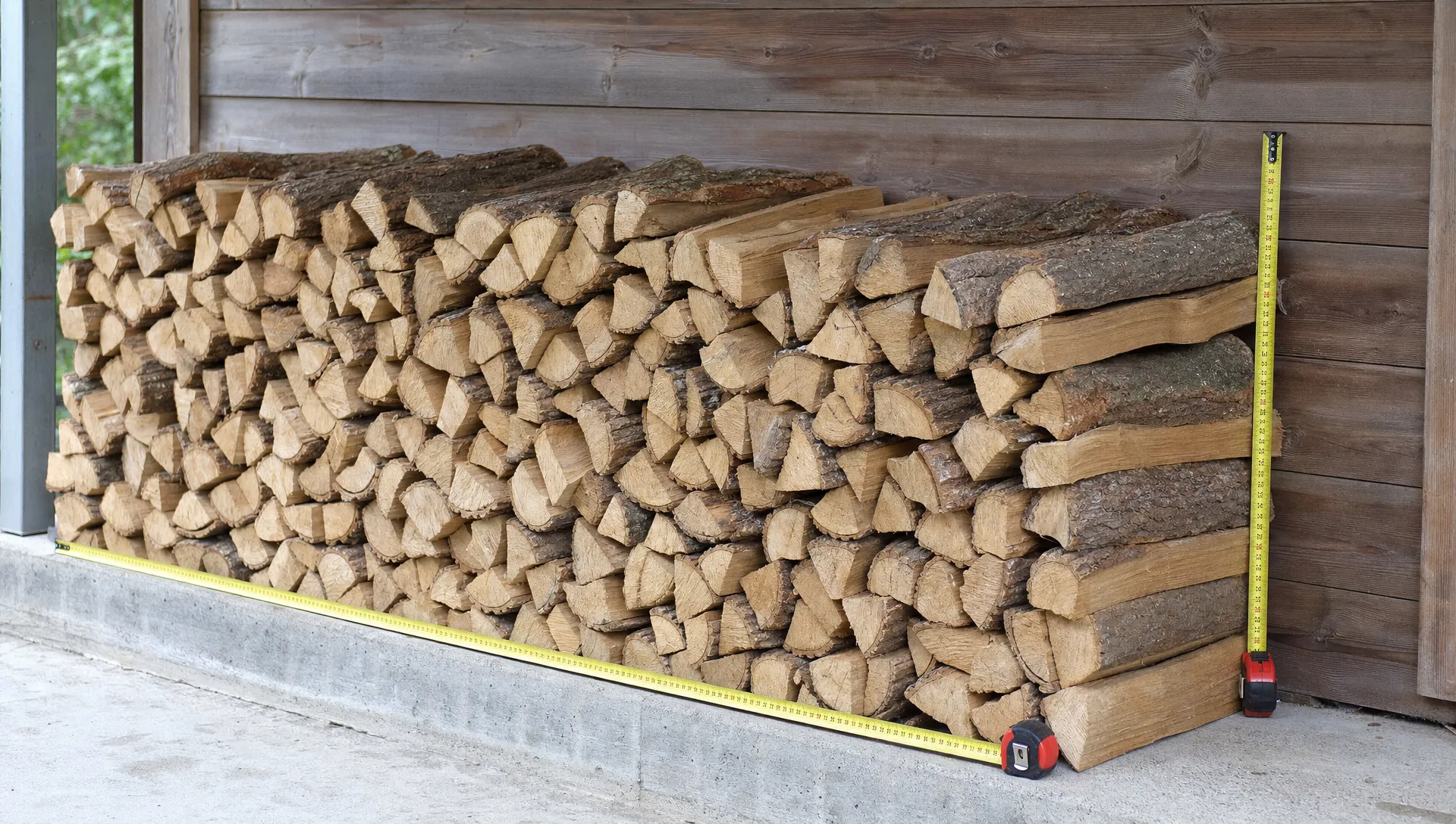 Une pile de bûches de chauffage rangée contre un mur d’abri, avec un mètre ruban posé le long de la face avant pour montrer la longueur des bûches et la hauteur de l’empilement.
